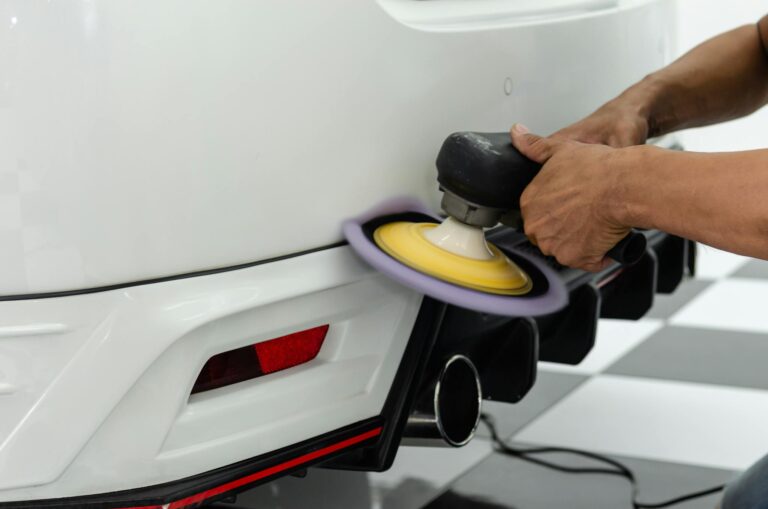 Detailer polishes a white car bumper with a power tool in a workshop setting.